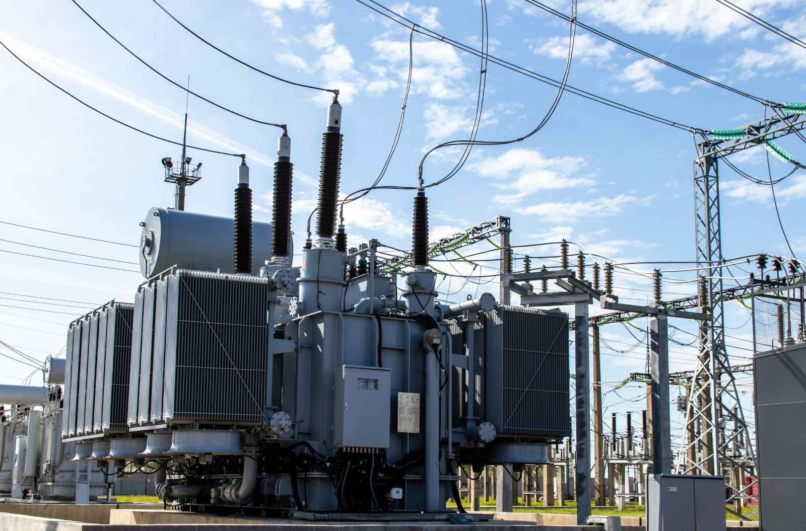 A large electrical transformer in a substation with cooling radiators, bushings, and a control panel, surrounded by transmission towers and power lines under a partly cloudy sky