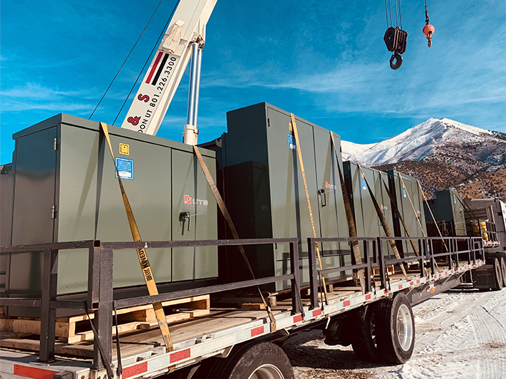 A flatbed truck with industrial cabinets is parked near snow-covered mountains under a clear blue sky, with a crane overhead.