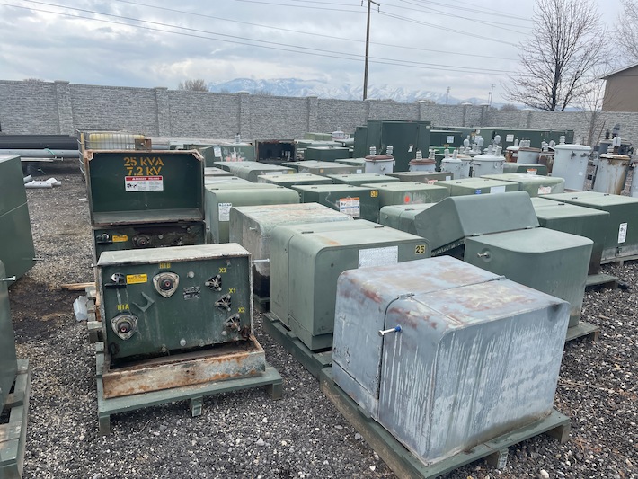 A storage yard with electrical transformers and switchgear, including a 25 kVA, 72.2 kV unit, under a cloudy sky with mountains in the background.