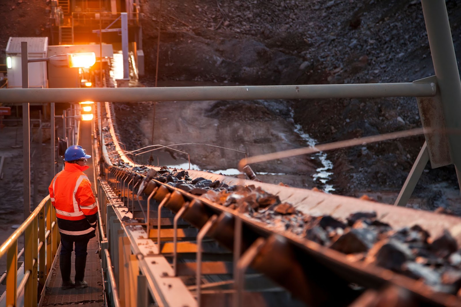 A worker in a hard hat and orange safety gear oversees a conveyor belt moving rocks in a mining facility.