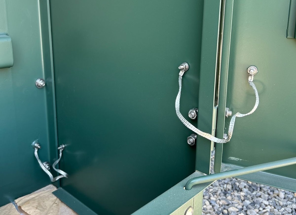 Green electrical cabinet with open door and metal straps,placed on a gravel surface.