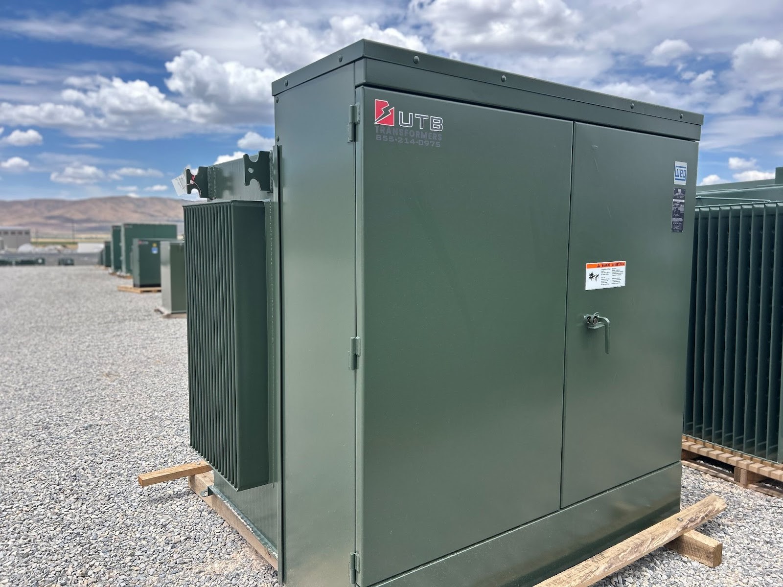 Green electrical transformer unit with UTB Transformer branding, placed on a gravel surface outdoors.