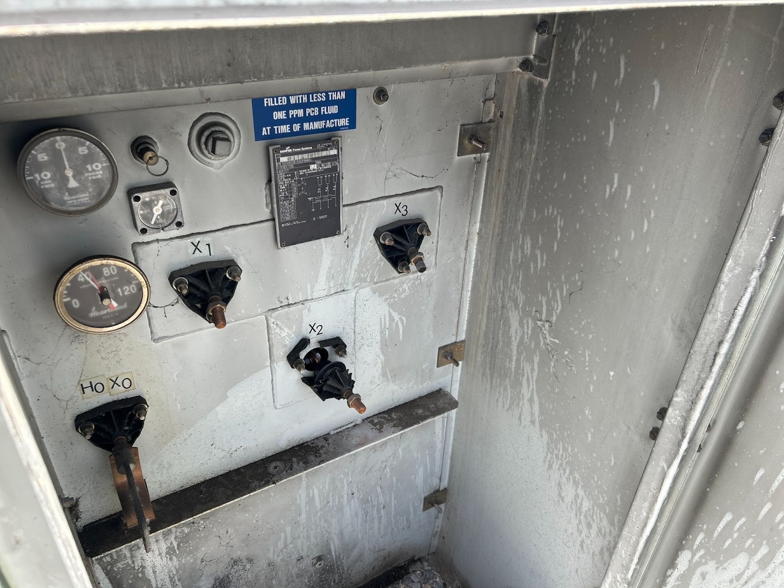 Interior view of a weathered transformer panel with gauges, connection points labeled X1, X2, X3, and Ho Xo, and a notice about PCB fluid.