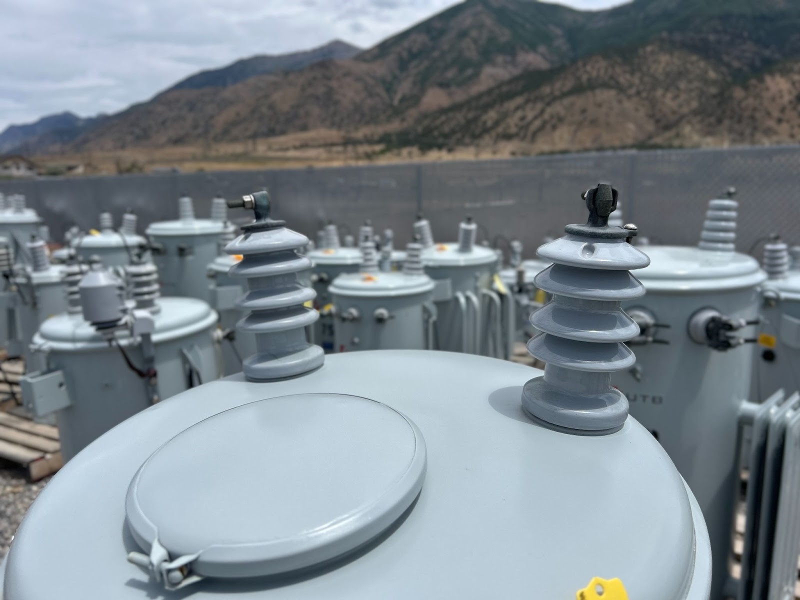 Outdoor transformer substation with high-voltage equipment and mountain landscape in the background.