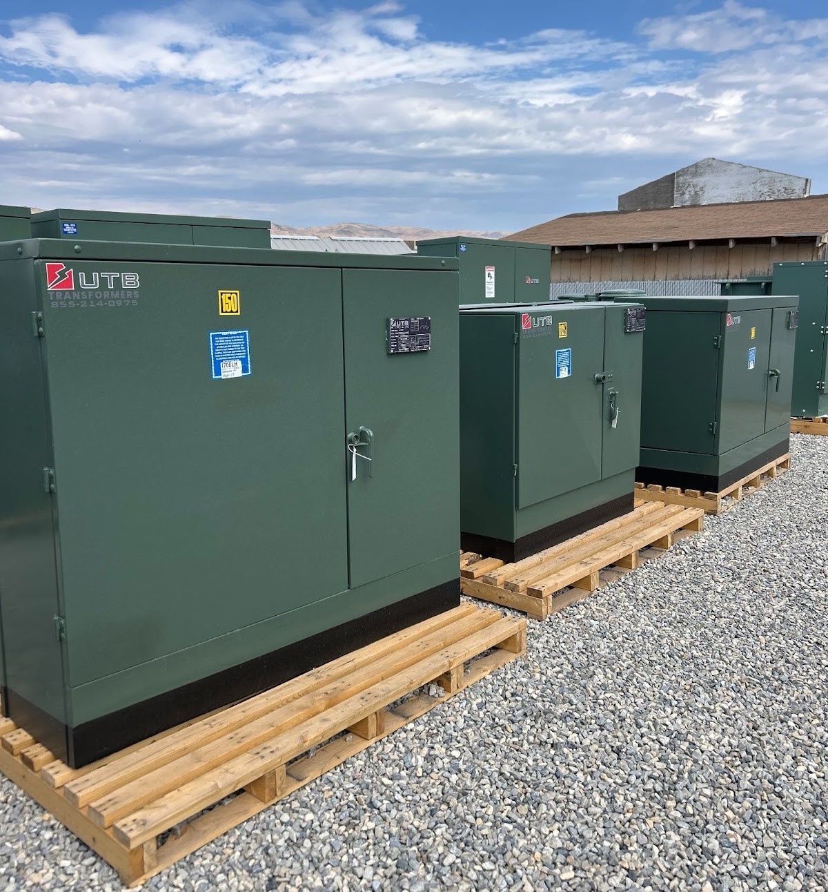 The image shows a row of green UTB Transformers cabinets labeled 150 on wooden pallets, placed on a gravel surface under a clear blue sky with mountains in the background.