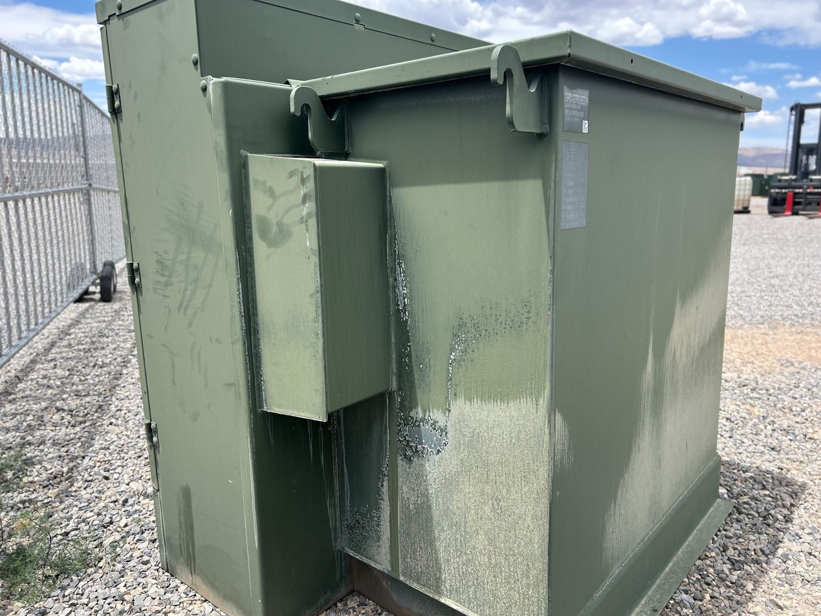 Weathered green transformer unit with hooks on top, placed on a gravel surface near a fence.